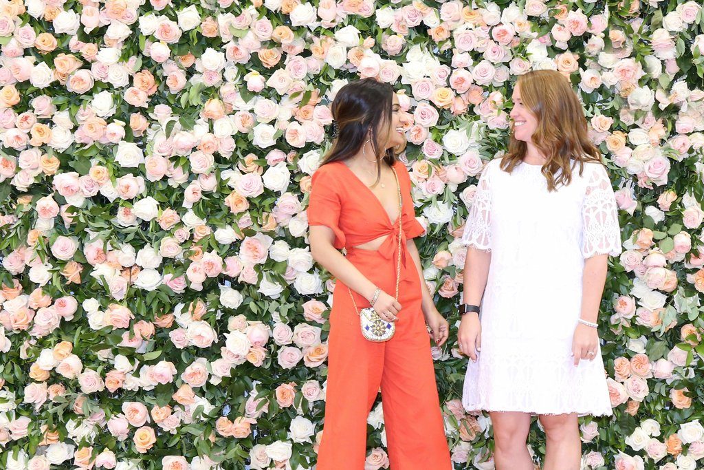 Pooja Gandhi and Shannon Fresca are photographed standing in front of a floral wall at Project Womens Las Vegas.