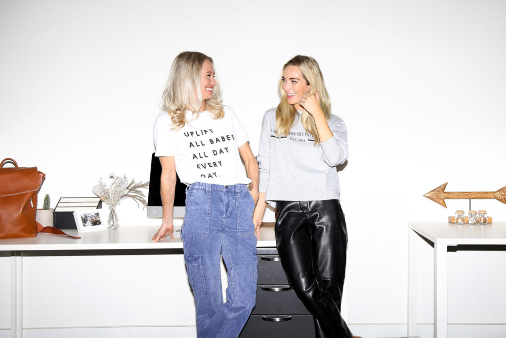 Female founders Sam Ellis and Hannah Bernard of Roots and Ardor are photographed standing and smiling.